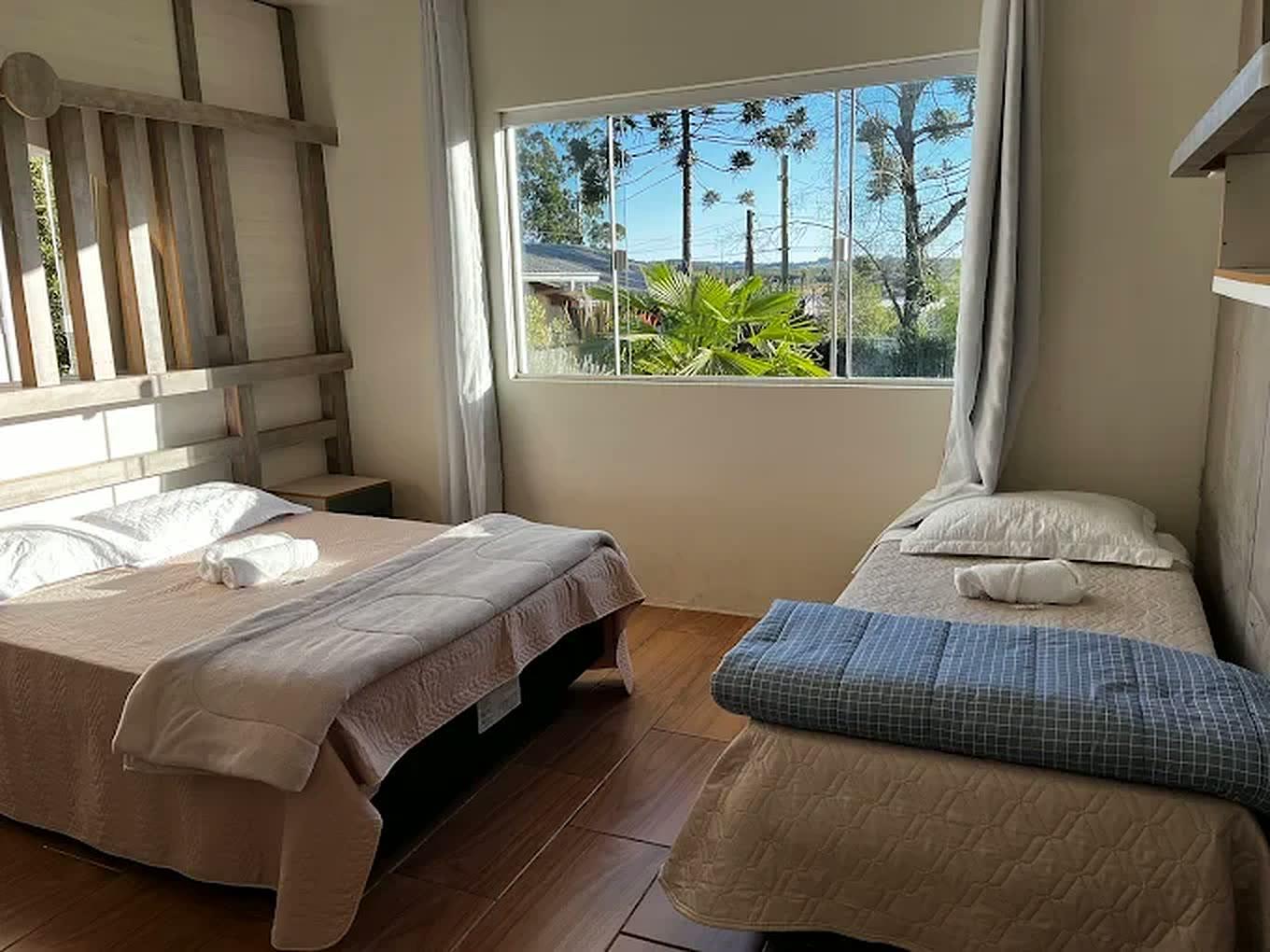 Quarto com cama de casal e cama de solteiro, janela ampla com araucárias ao fundo